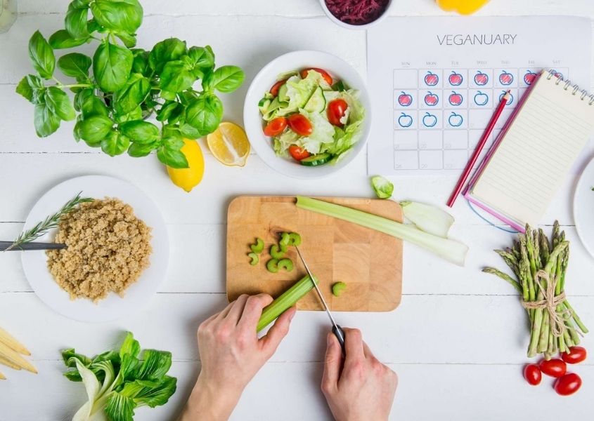 White table with women's hands chopping vegetables on wooden board with various vegetables on table