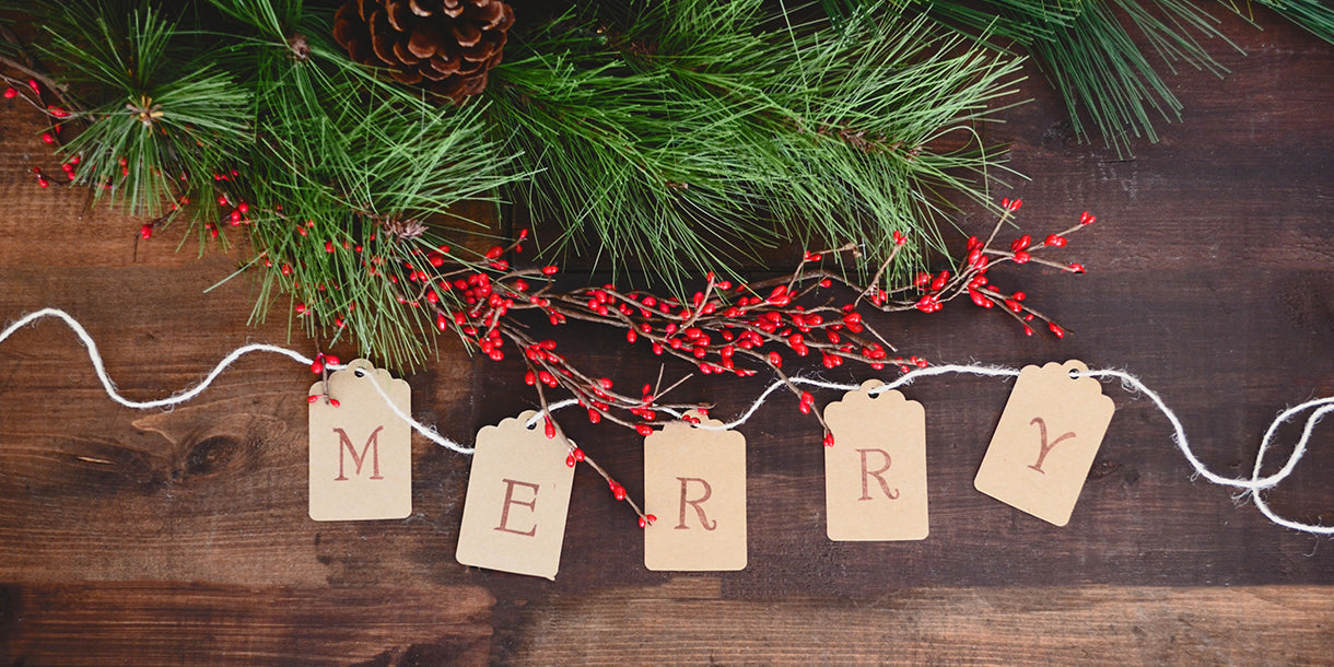 Gift tags spelling out 'merry' on a wooden surface with Christmas berries and green twigs