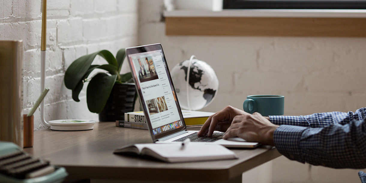 A man sitting by a wooden desk typing on a laptop with a green plant in the background