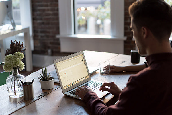 Man sitting at a dark wooden desk with a laptop and a cluster of houseplants and flowers