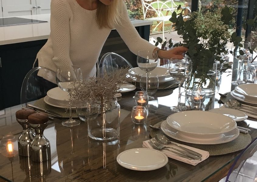 Woman wearing white jumper leaning over a reclaimed wood dining table with glasses, plates and flowers