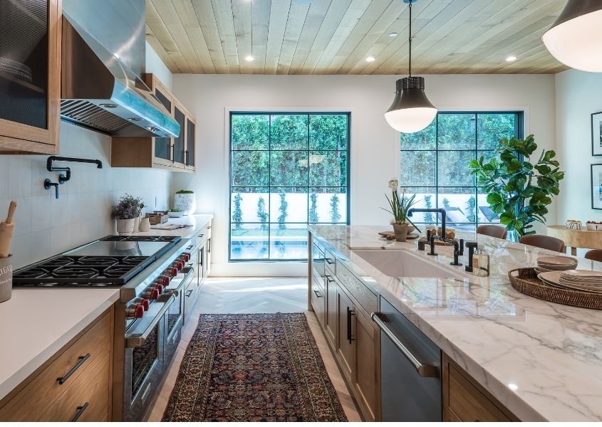 View of a kitchen extension with two large windows and a galley kitchen with wood units and  a marble top