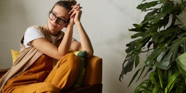 Woman dressed in mustard trousers resting on an armchair next to green plant