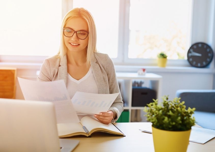 Blonde lady sat at desk with laptop and green plant in front of a large bright window
