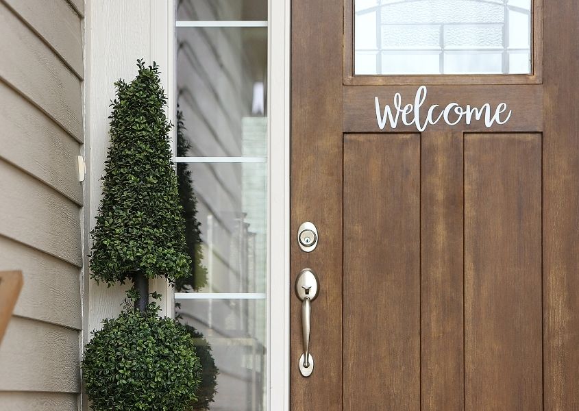 Natural wooden front door with words welcome on front with green topiary at entrance