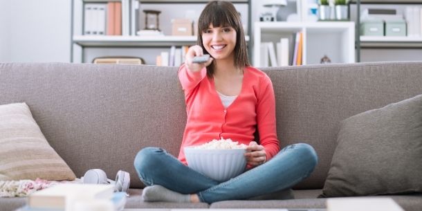 Girl in pink cardigan sat on sofa with bowl of popcorn holding tv remote control